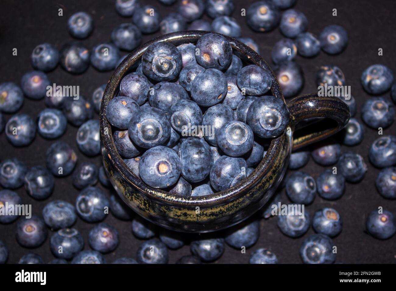 Una tazza di ceramica nera, riempita a più di versamento con grandi mirtilli color indaco, con più bacche blu che la circondano Foto Stock