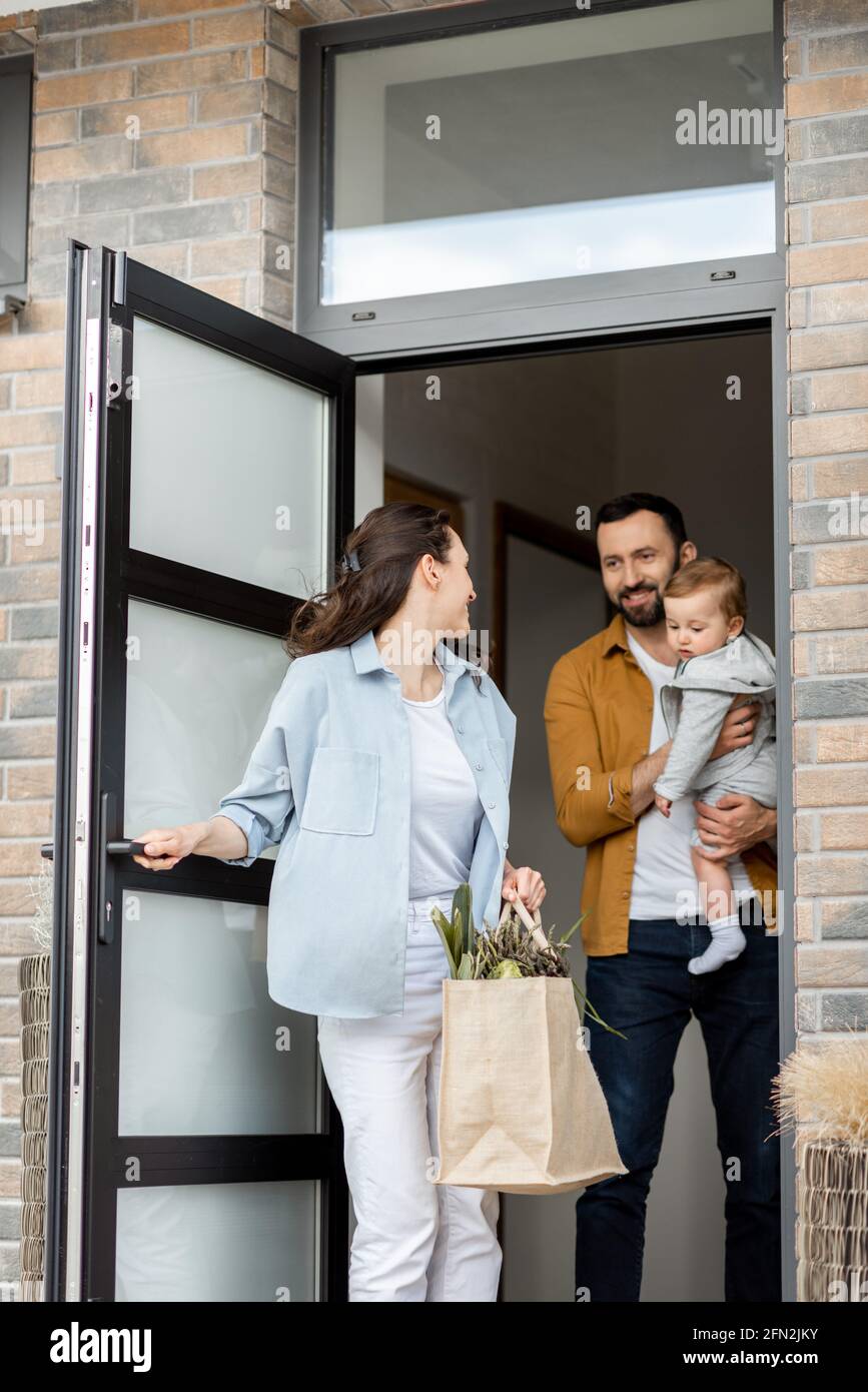 I giovani genitori con il bambino neonato ordinano la consegna del cibo online e si trovano di fronte alla casa per riceverlo. Casalinga porta la borsa della spesa mentre il marito tiene un bambino. Foto Stock
