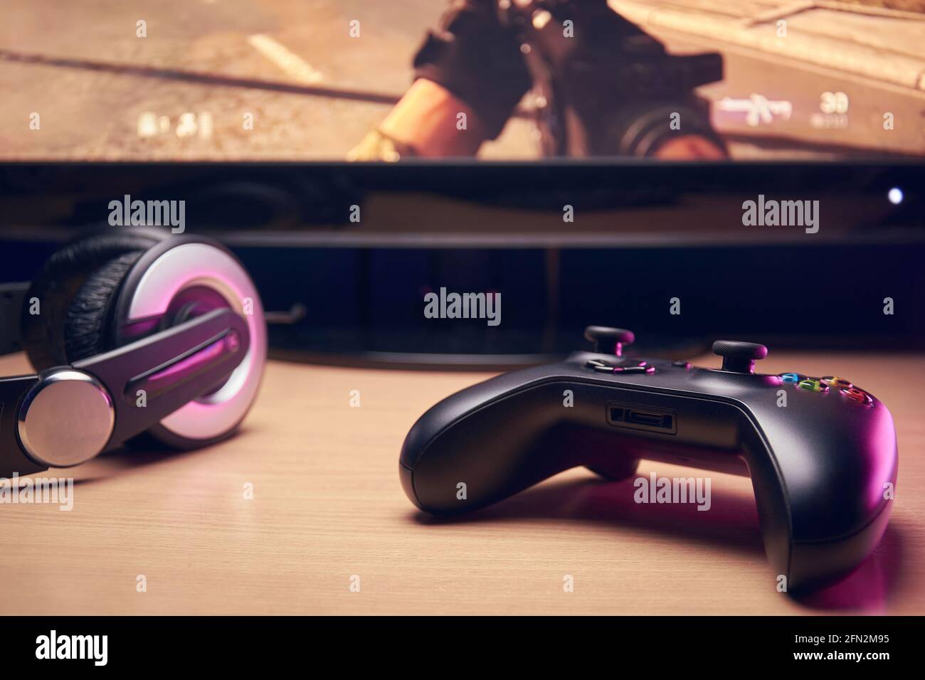 Visualizzazione della configurazione della scrivania di gioco con il gamepad nero illuminato da luci viola, cuffie e schermo che mostrano l'interfaccia di gioco video. Foto Stock
