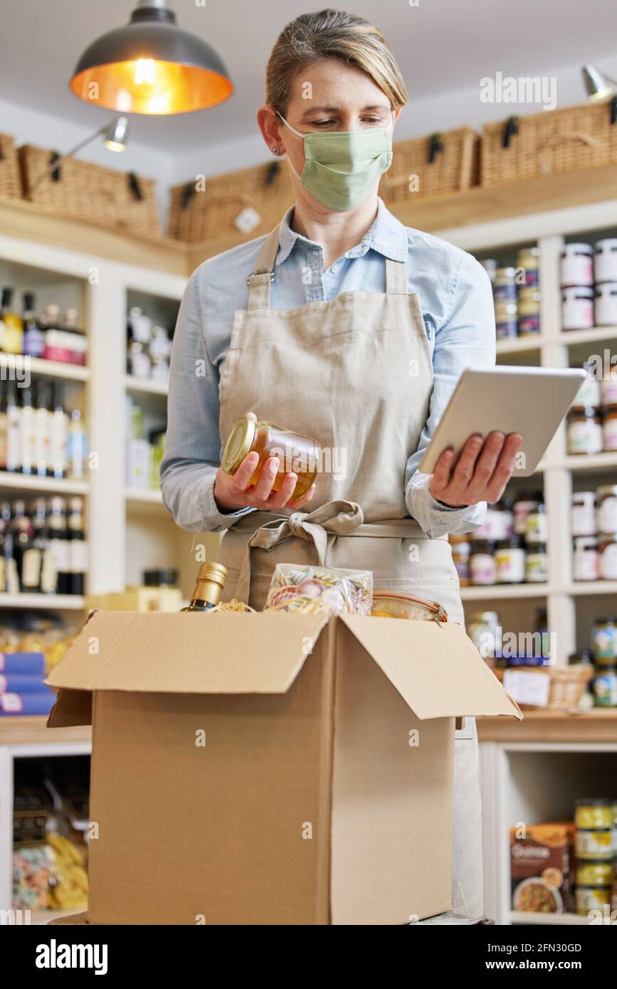 Donna Proprietario Delicatessen con Tablet digitale indossare maschera di preparazione Ordine di alimentari online Foto Stock