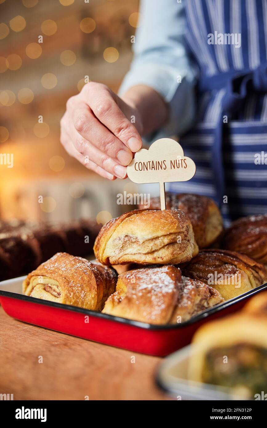 Sales Assistant in panetteria che mette contiene l'etichetta NUTS nella pila Di coniglietti di cannella appena sfornati Foto Stock
