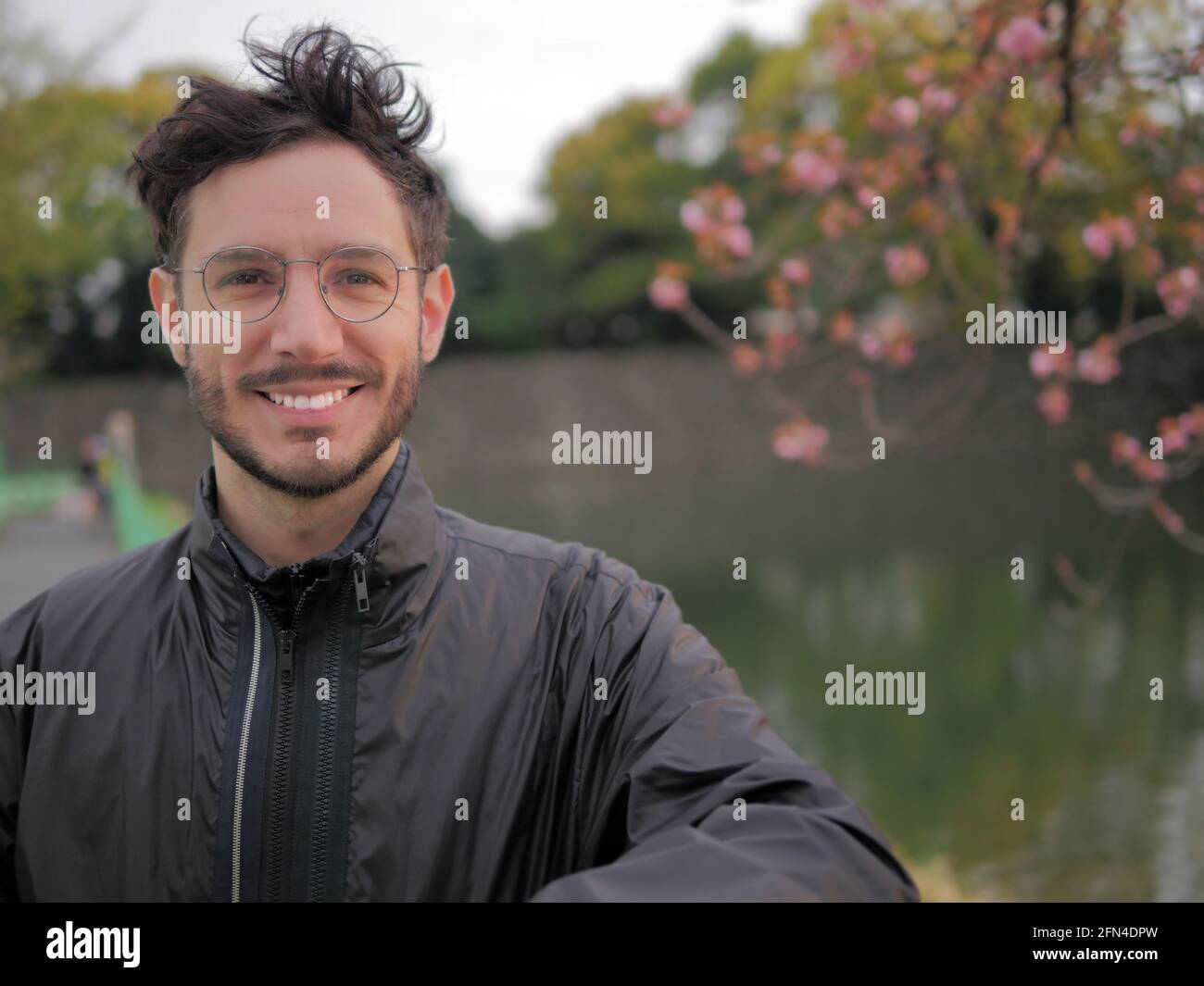 Un uomo sorridente con gli occhiali si pone davanti a un lago giapponese. Turisti che visitano Tokyo in primavera. Uomo carino e ciliegi in fiore. Foto Stock