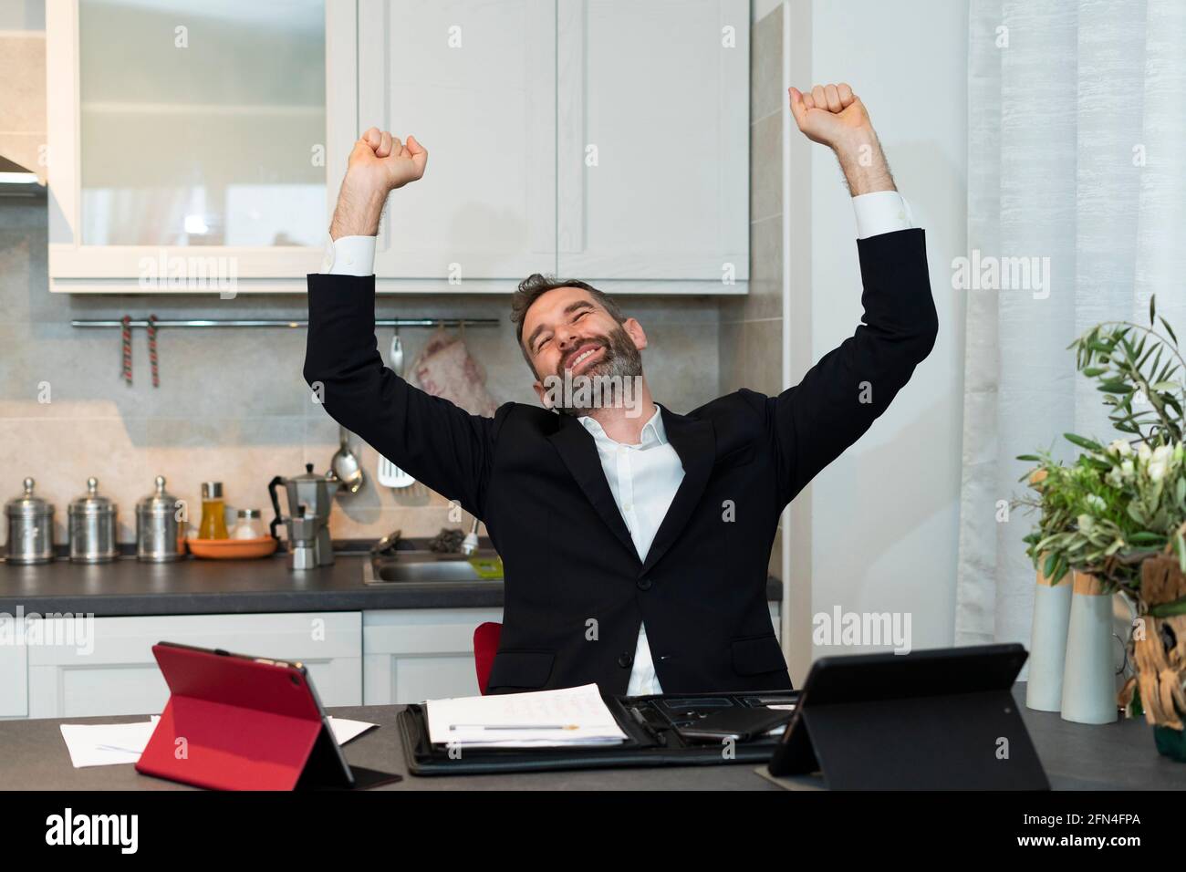 Ritratto di gioioso giovane uomo d'affari che alza entrambe le braccia per un affare all'alba. L'operaio domestico espressivo gioisce nella cucina. Riunione di successo. Foto Stock