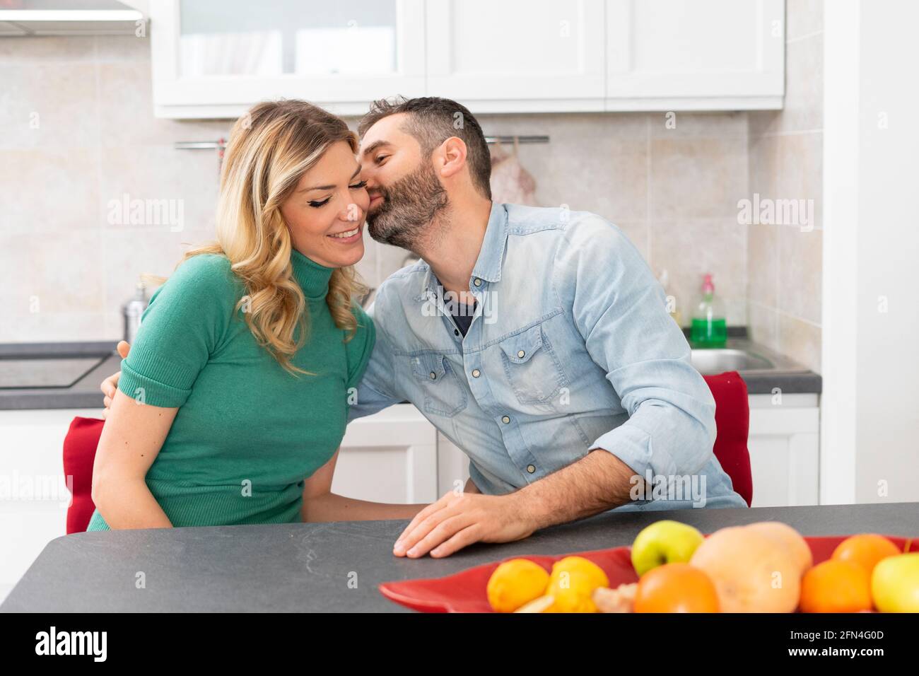 La coppia felice sta baciando in cucina. Sposi seduti al tavolo da cucina. Il ragazzo bacia la sua ragazza bionda. Buon bacio del mattino. Foto Stock