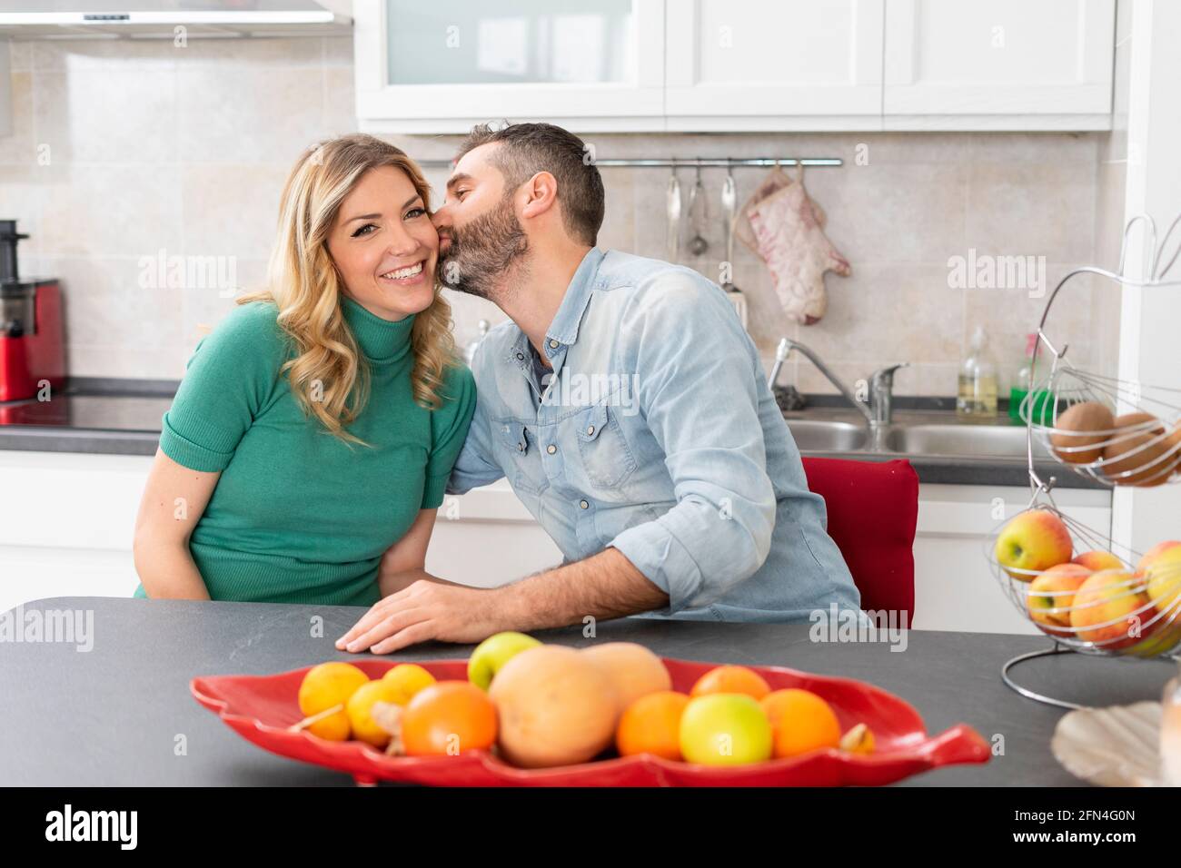 La coppia felice sta baciando in cucina. Sposi seduti al tavolo da cucina. Il ragazzo bacia la sua ragazza bionda. Buon bacio del mattino. Foto Stock