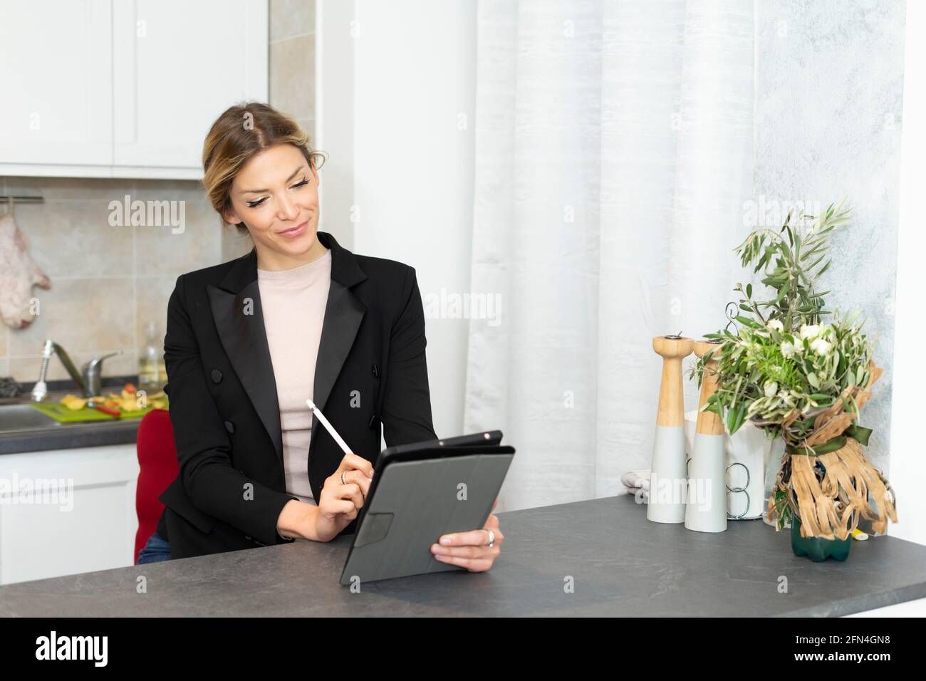 Attraente donna di affari lavora da casa. Elegante ragazza in tuta nera con tablet e prendendo appunti sul tavolo da cucina. Lavoro intelligente in blocco. Foto Stock