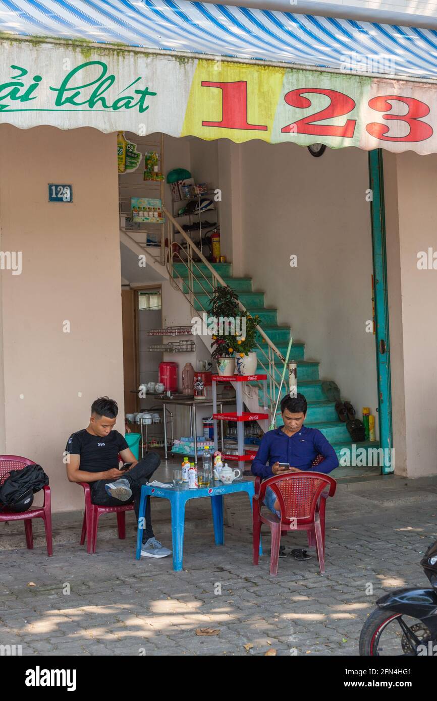 Due vietnamiti seduti agghiaccianti fissando i loro telefoni cellulari all'esterno della caffetteria sul pavimento, da Nang, Vietnam Foto Stock