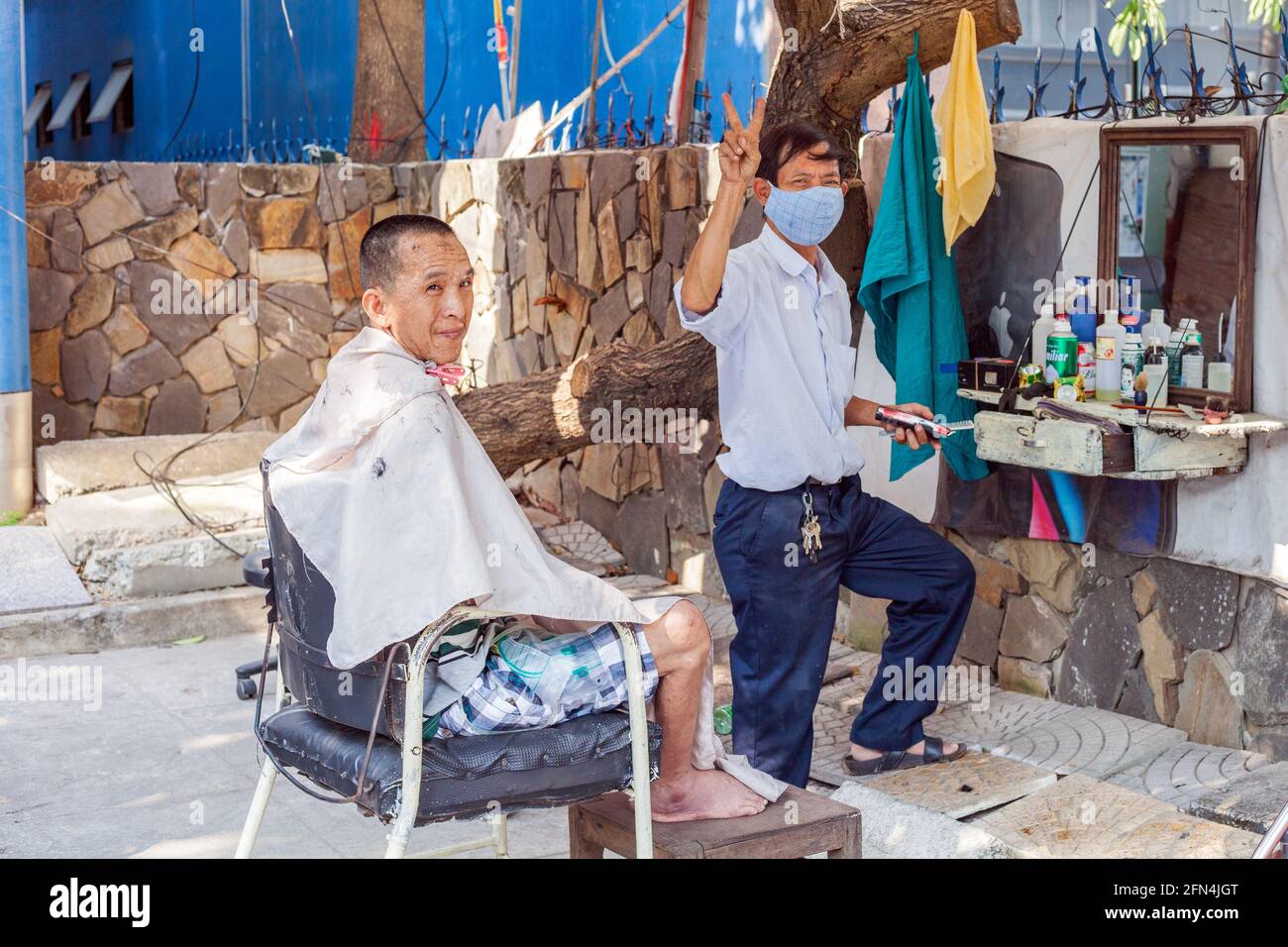 Vietnamita barbiere di strada dà V segno come lui taglia i capelli del maschio, da Nang, Vietnam Foto Stock