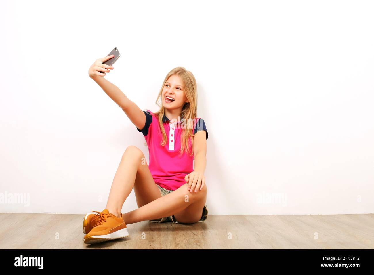 Bella ragazza teenage seduta su pavimento di legno che prende selfie colpi sul suo telefono delle cellule. Casual giovane femmina in sneakers giallo si fotografa su SM Foto Stock