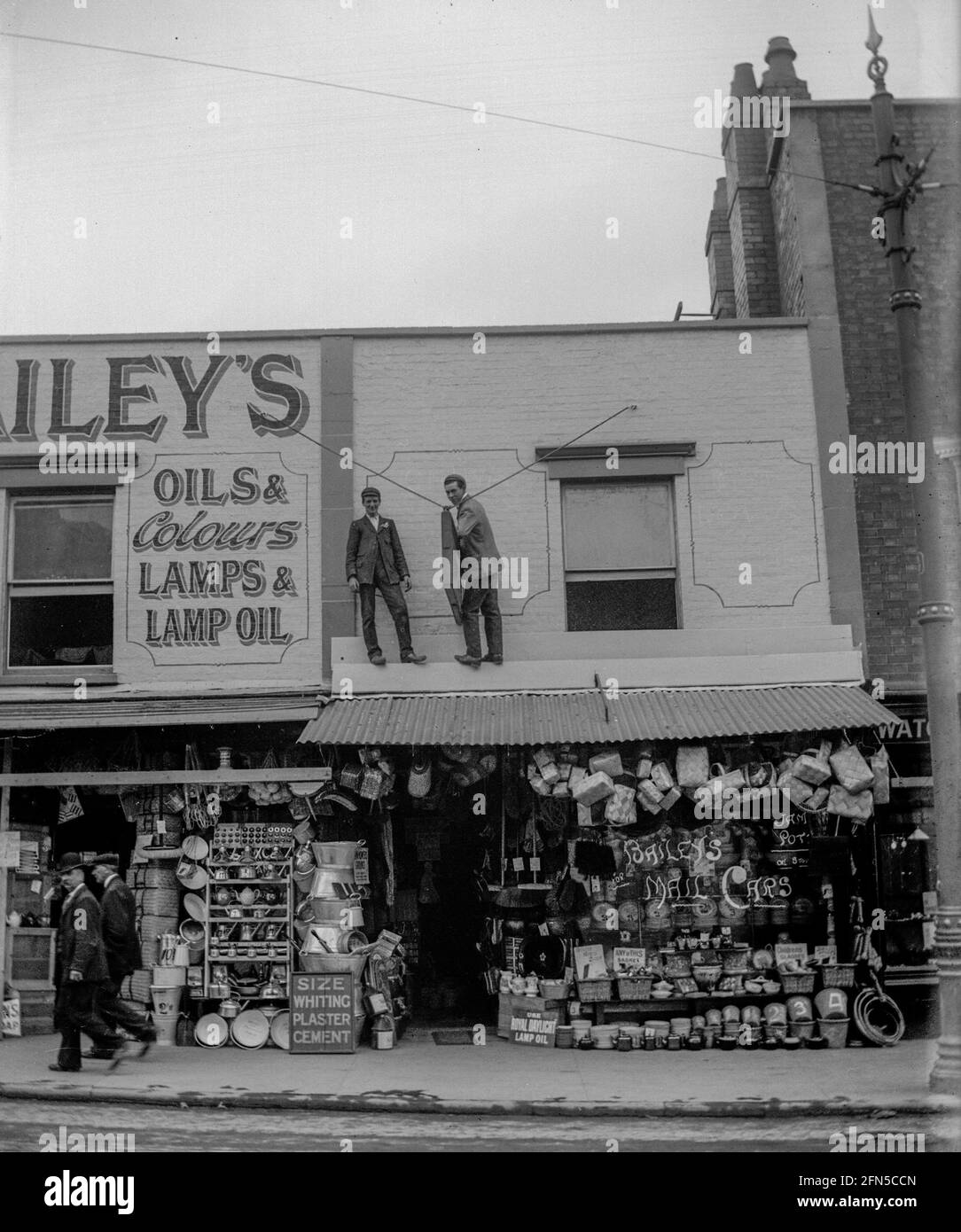 Due uomini lavorano sulla tettoia dei negozi Bailey's a Gloucester Road, Bristol intorno al 1910. Un negozio di ferramenta con le sue scorte molto in mostra dalla strada nei giorni in cui l'olio lampada era importante per tutti. Foto Stock