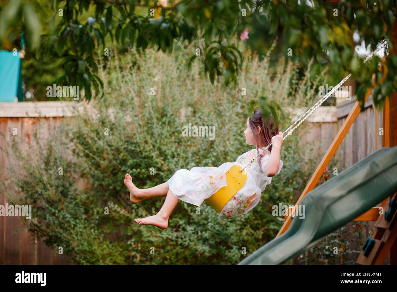 Una ragazza felice a piedi nudi oscilla su un playlet sotto a. albero in estate Foto Stock