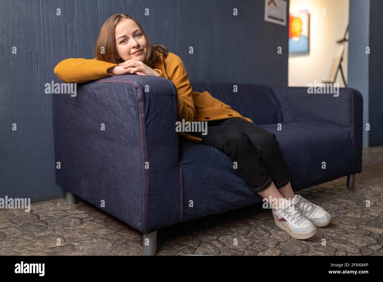 Ritratto di una giovane adolescente poggiando la testa sulle mani sul retro del divano. Giallo su sfondo blu. Foto Stock