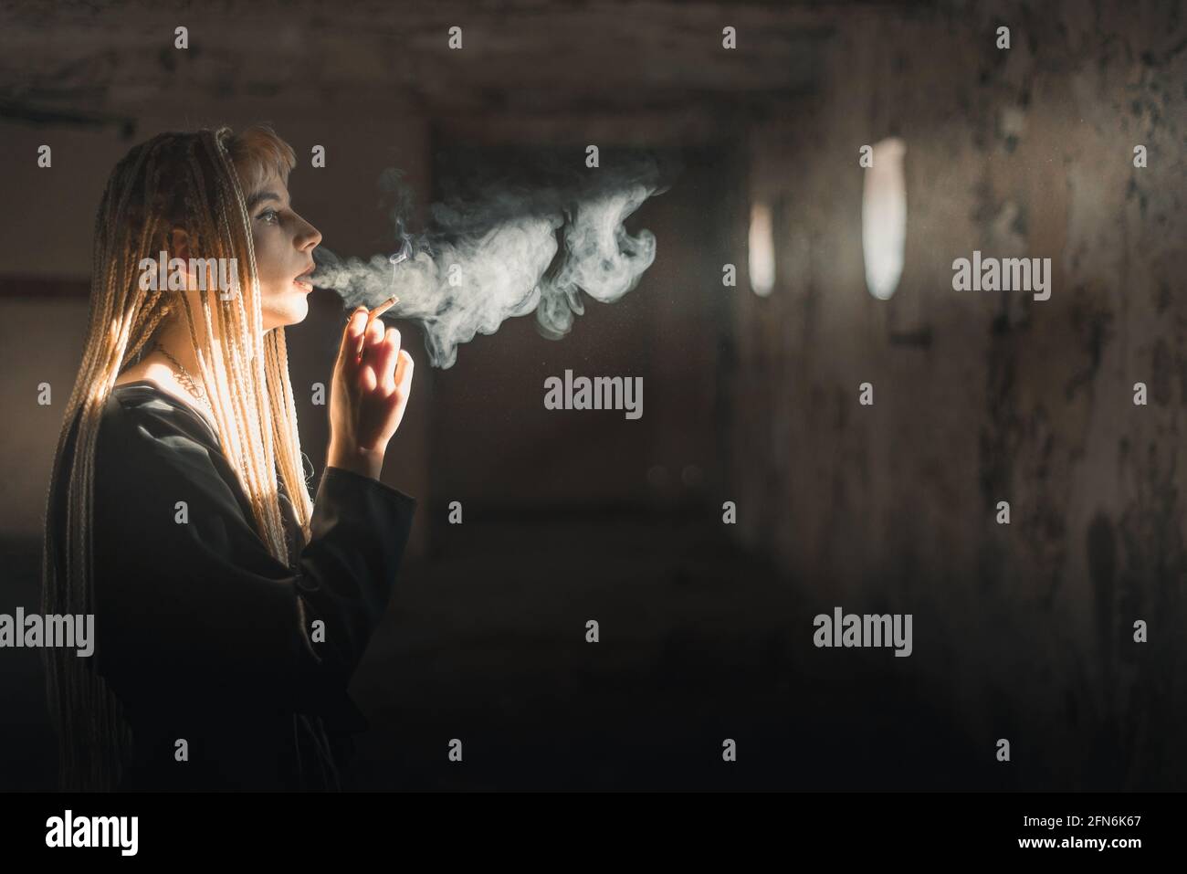 Giovane donna bionda che fuma una sigaretta in camera rovinata con luce che si illumina dalla finestra Foto Stock
