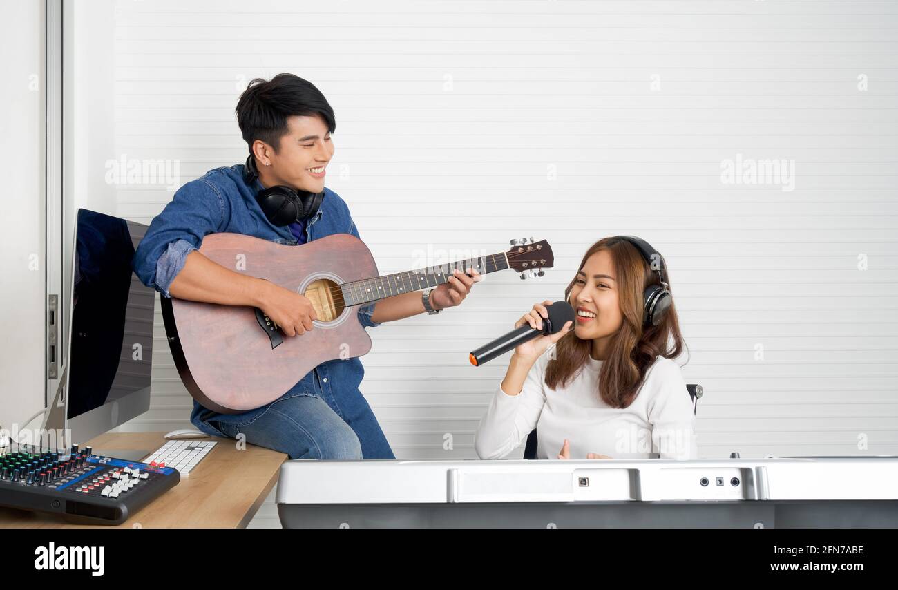 Giovane donna asiatica cantare mentre si gioca una tastiera elettrica di fronte alla parete bianca. Il suo ragazzo suona la chitarra insieme. Musicisti che producono musica in pro Foto Stock