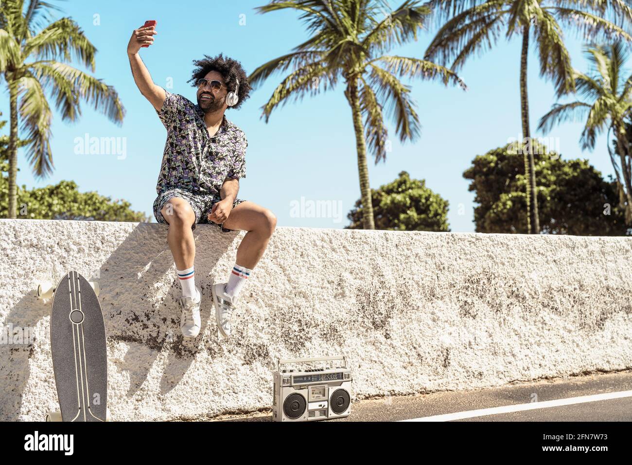 Giovane latino che si diverte a prendere selfie con smartphone mobile mentre ascolti la musica con le cuffie e il stereo portatile durante le vacanze estive Foto Stock