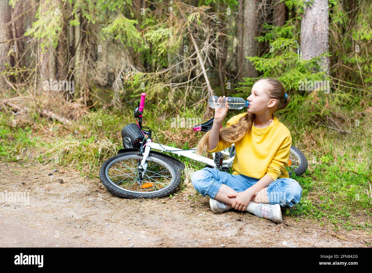 Una ragazza con una bicicletta si siede su un sentiero in un parco o in una foresta, beve acqua da una bottiglia. Foto Stock