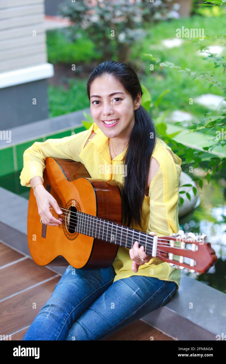 Buon musicista sorridente e divertente godere hobby concetto. Donna asiatica che suona la chitarra classica in cortile, all'aperto con luce solare intensa. Ronzio positivo Foto Stock