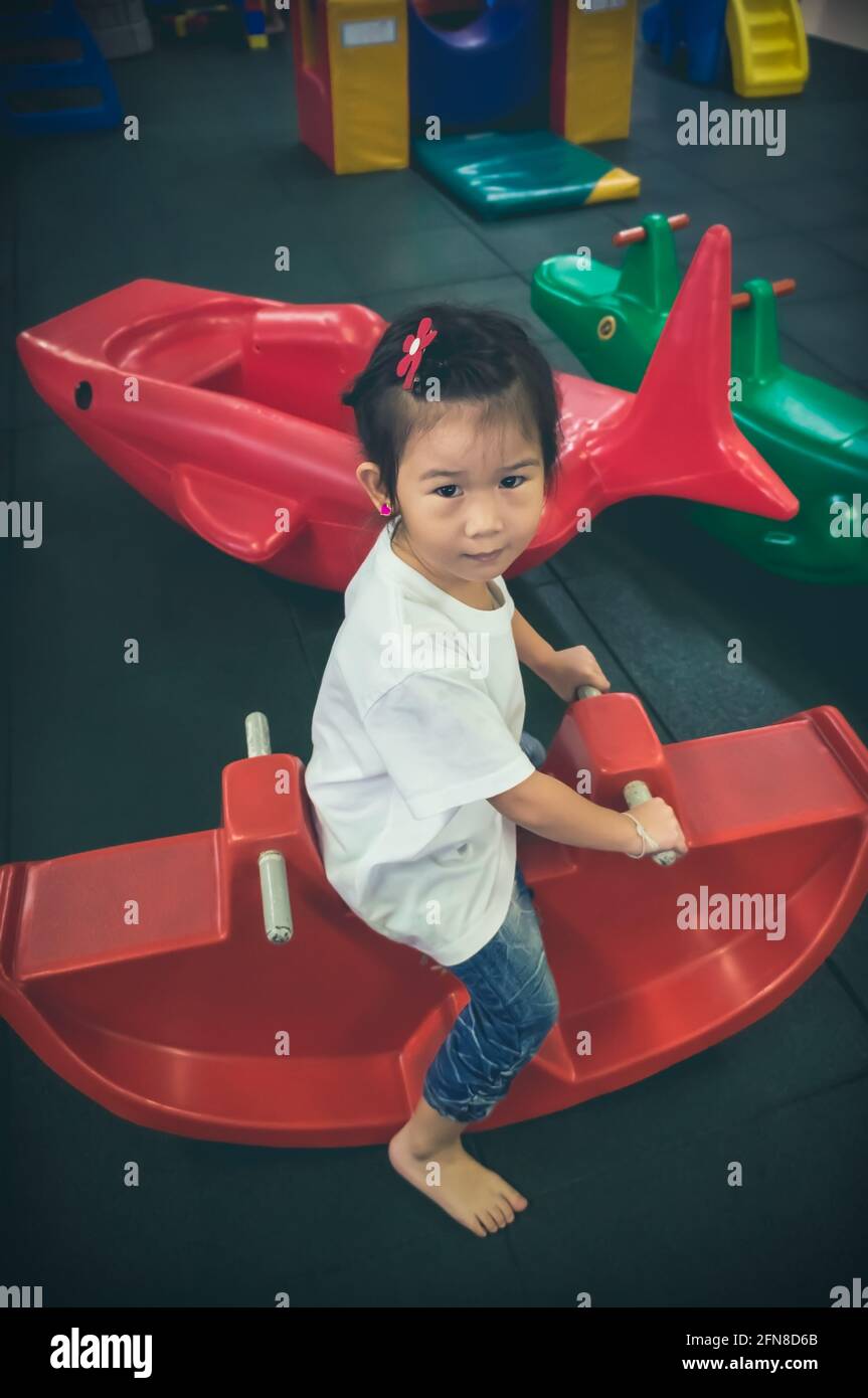 Carino bambino asiatico divertirsi e guardare la macchina fotografica mentre si sente felice al parco giochi per bambini al coperto. Ragazza asiatica seduta su una sega di plastica rossa. Vintage Foto Stock