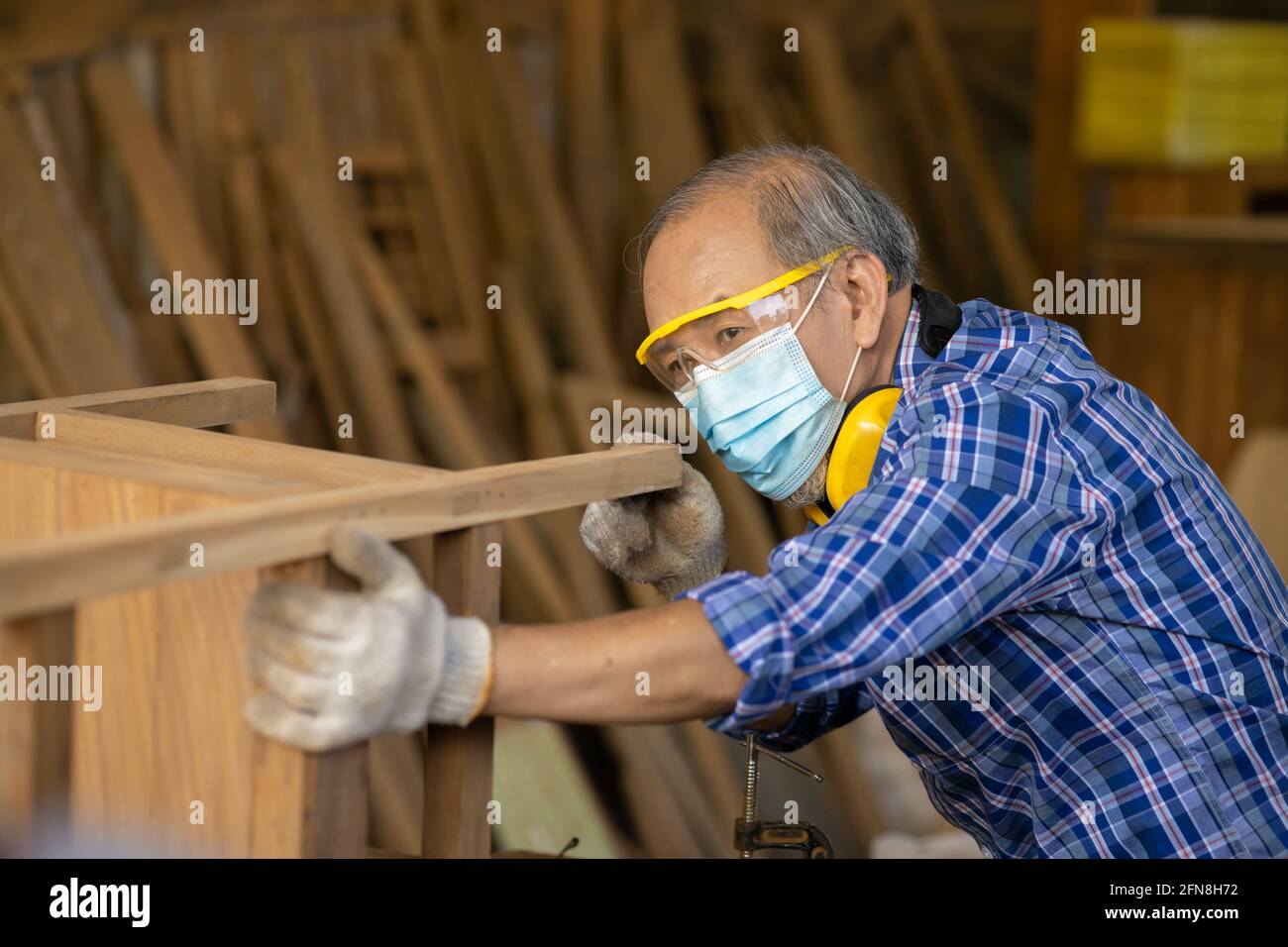 Anziano lavoratore legno woodcraft ritire hobby per il buon pensionamento, asiatico maschio maturo maestro professionale di fare mobili in legno con maschera protects viso Foto Stock
