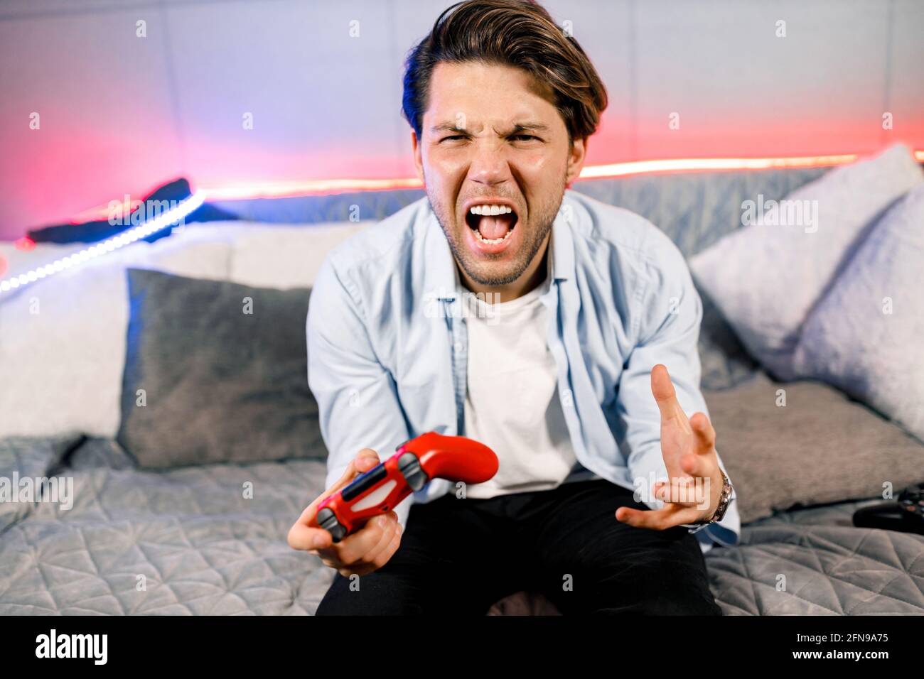 Foto di bel ragazzo eccitato seduto su un comodo divano, quarantena rimanere a casa giocatore tenendo in mano il joystick stazione di gioco, buon umore campionato fifa, infelice Foto Stock