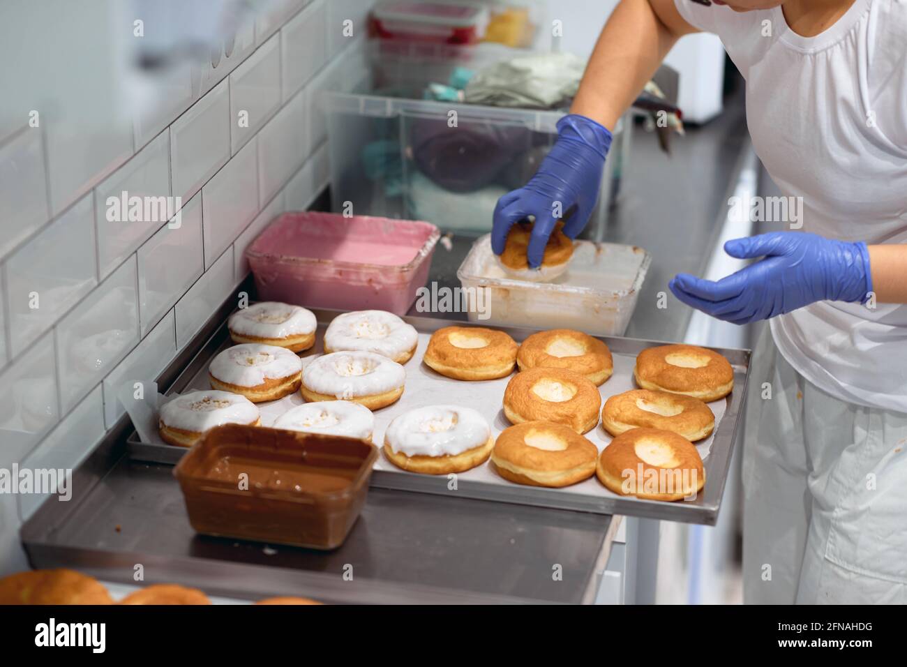 Una lavoratrice cede le ciambelle in un delizioso condimento in un'atmosfera di lavoro in un laboratorio di caramelle. Pasticceria, dessert, dolce, fare Foto Stock