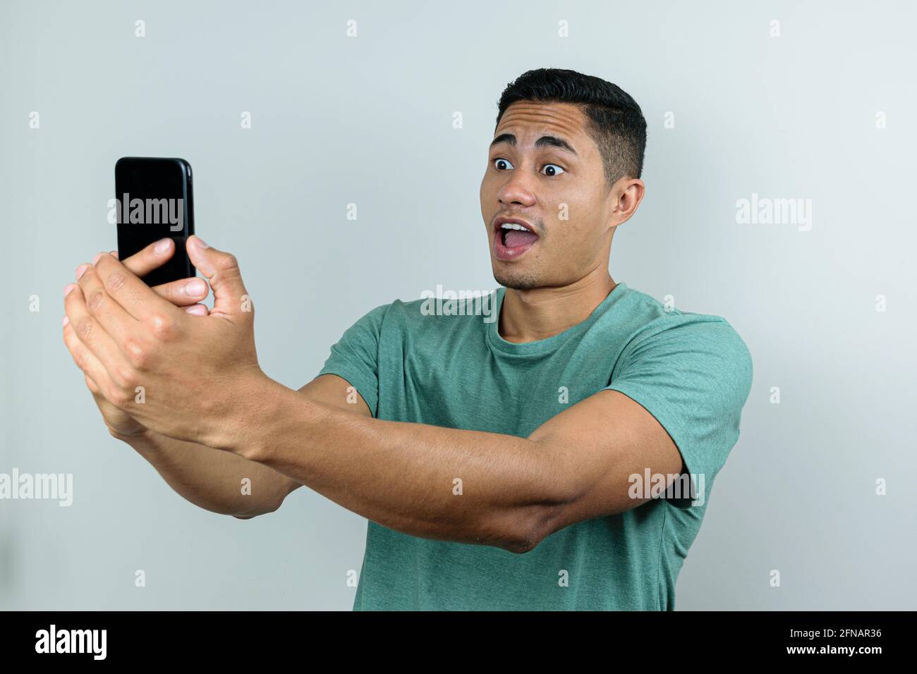 Giovane brasiliano di 28 anni, tenendo e guardando lo smartphone, con gli occhi spalancati e la bocca aperta (sorpreso). Foto Stock
