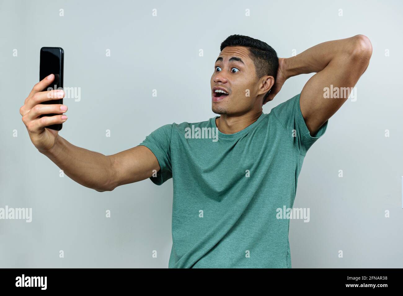 Giovane brasiliano, tenendo e guardando lo smartphone, con gli occhi spalancati e l'altra mano sulla schiena del collo (sorpreso). Foto Stock