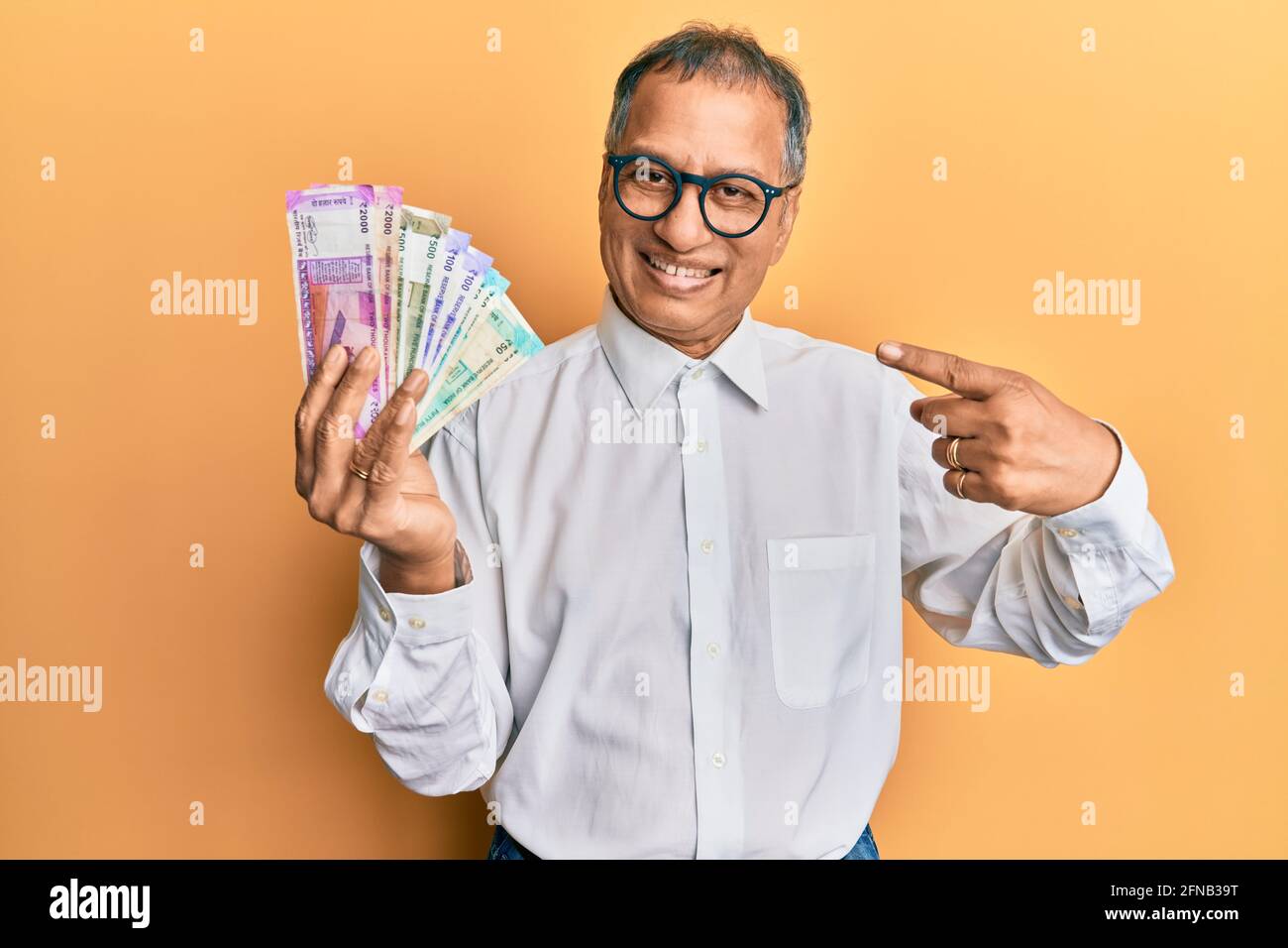 Uomo indiano di mezza età che tiene le banconote della rupia indiana sorridente felice puntare con la mano e il dito Foto Stock