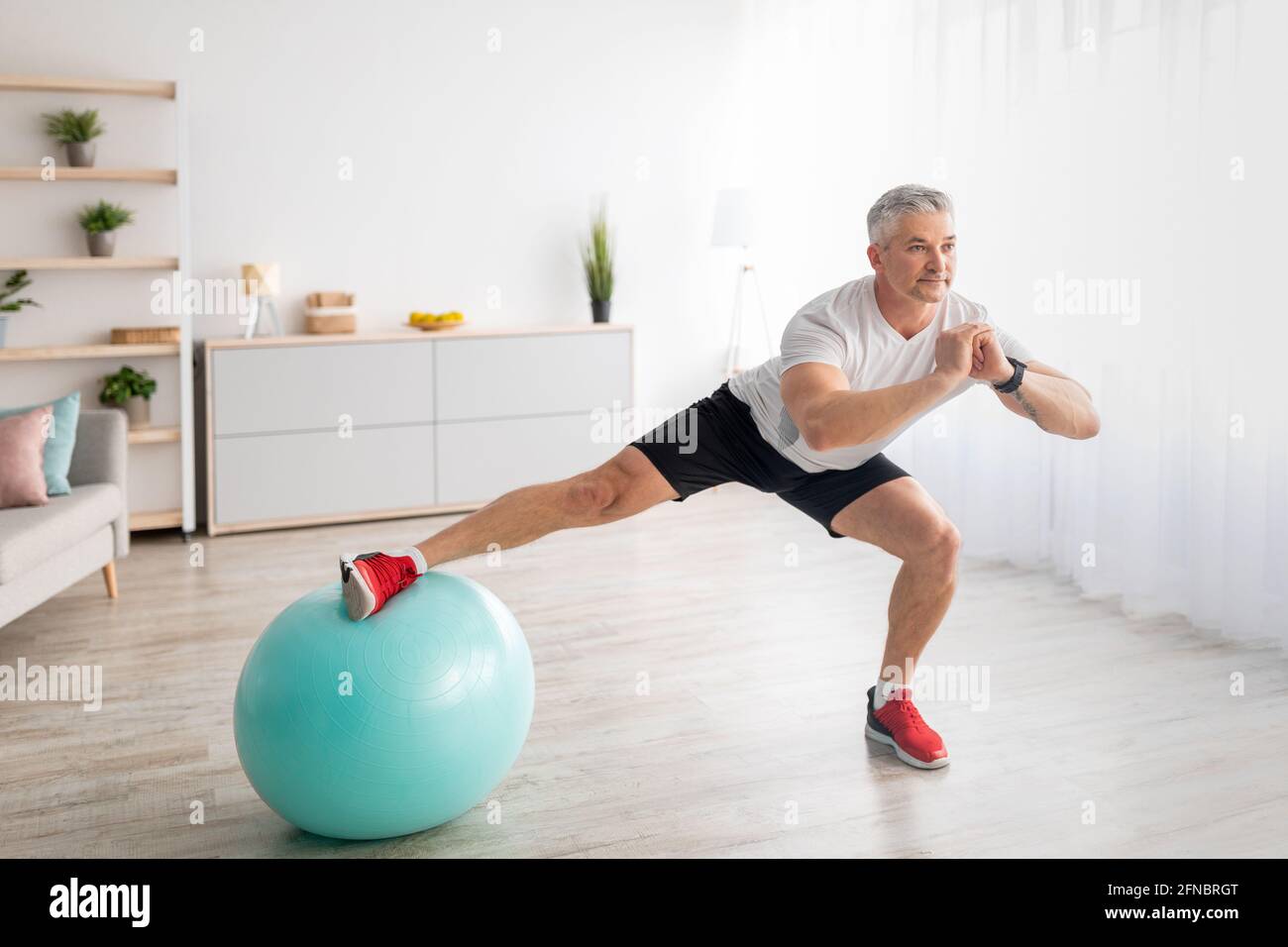 L'uomo maturo atletico che fa affondo laterale, usando la sfera di idoneità, che lavora fuori a casa nell'interno della stanza vivente Foto Stock