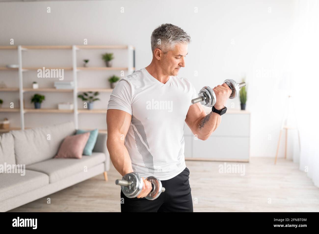 Uomo forte maturo che fa workout dumbbell a casa, lavorando sulla forza delle braccia e guardando i suoi bicipiti Foto Stock