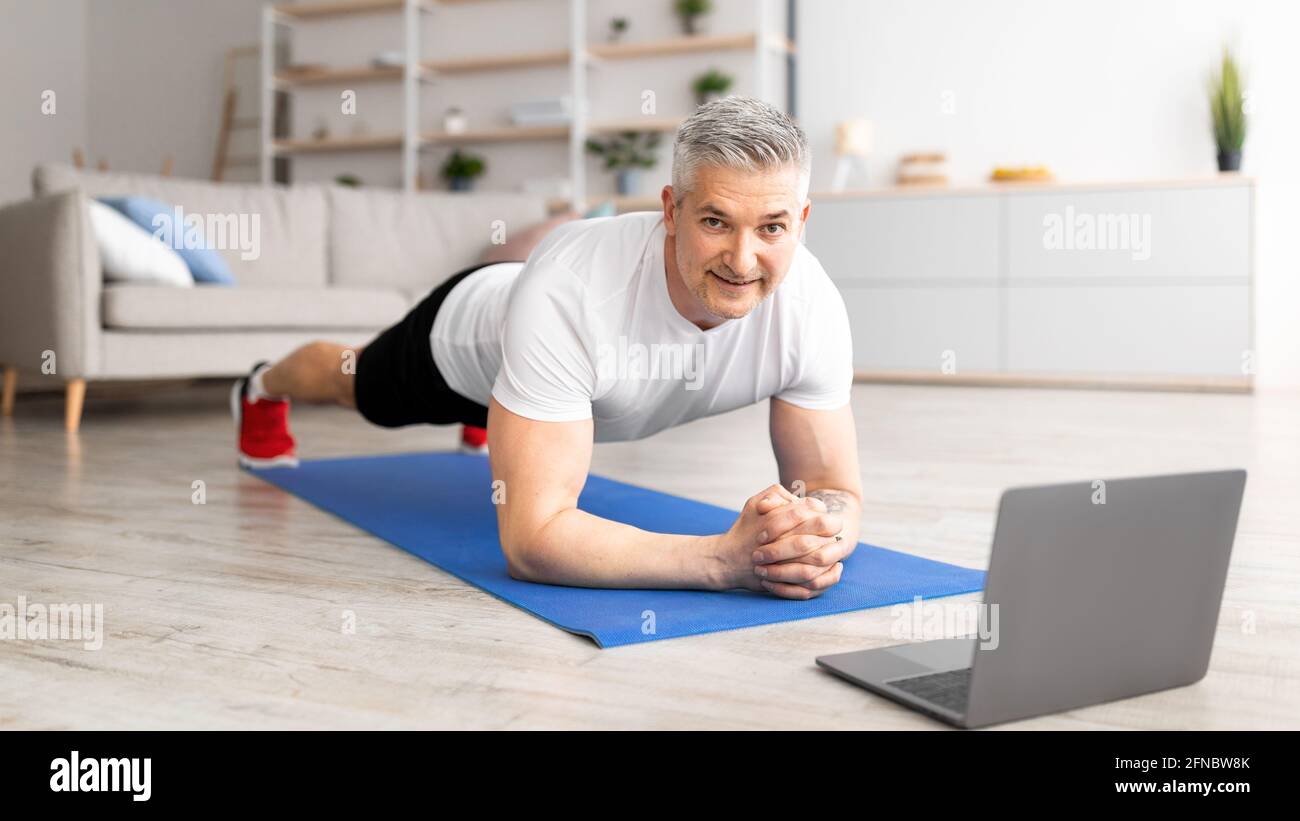Allenamento mattutino. Uomo sportivo di mezza età che fa la tavola di yoga, guardando il tutorial online sul computer portatile, allenandosi in salotto Foto Stock
