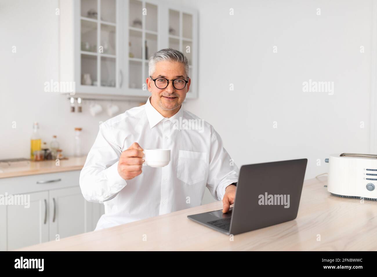 Tempo per il caffè del mattino. Uomo d'affari maturo che lavora su computer portatile e beve bevande calde, seduto in cucina Foto Stock