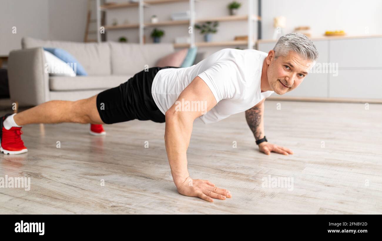 Routine di allenamento per il fitness maschile. Felice uomo anziano che fa spinta-in su sul pavimento, addestrando in soggiorno a casa, panorama Foto Stock