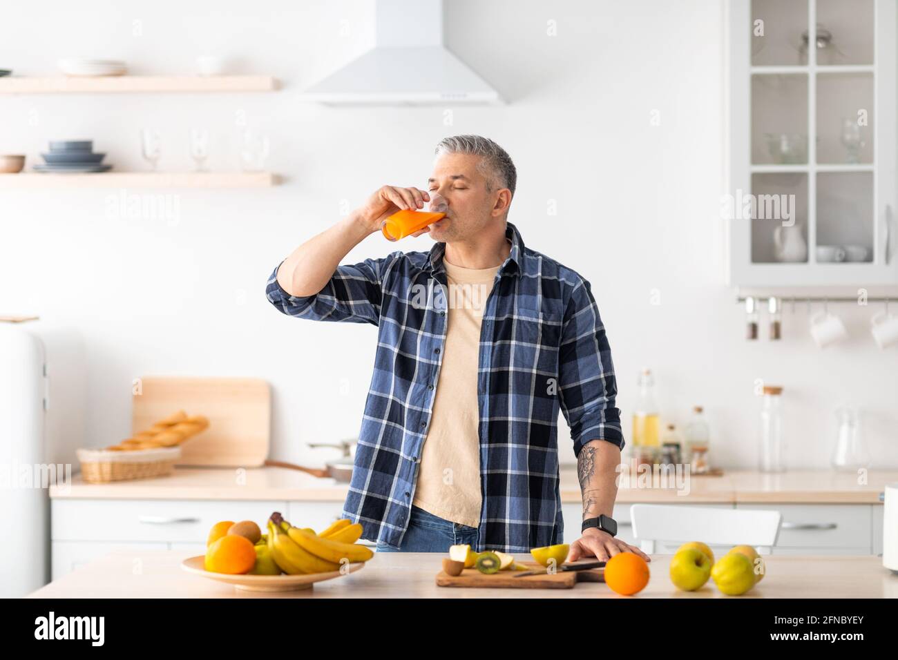 Mangiare sano. Uomo maturo che beve succo fresco, in piedi vicino al tavolo pieno di vari frutti in cucina interno Foto Stock