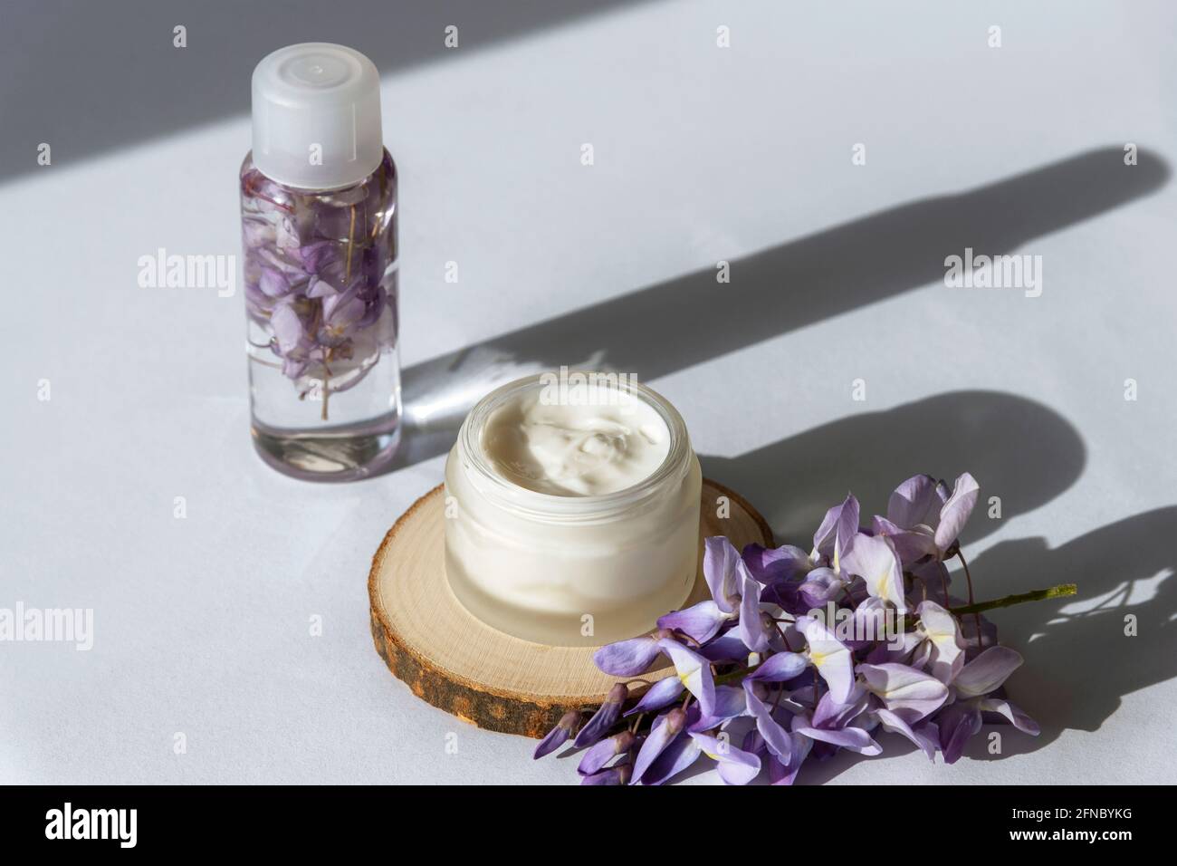 Vasetto di crema e fiori di glicine viola su tavola bianca alla luce del sole. Concetto di cosmetica biologica naturale. Lunghe ombre dure, vista dall'alto Foto Stock