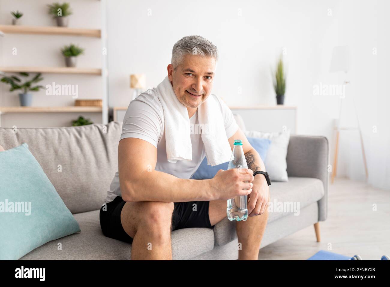 Uomo anziano sportivo che beve acqua fresca dopo l'allenamento sportivo a casa, seduto sul divano e sorridente alla macchina fotografica, copia spazio Foto Stock