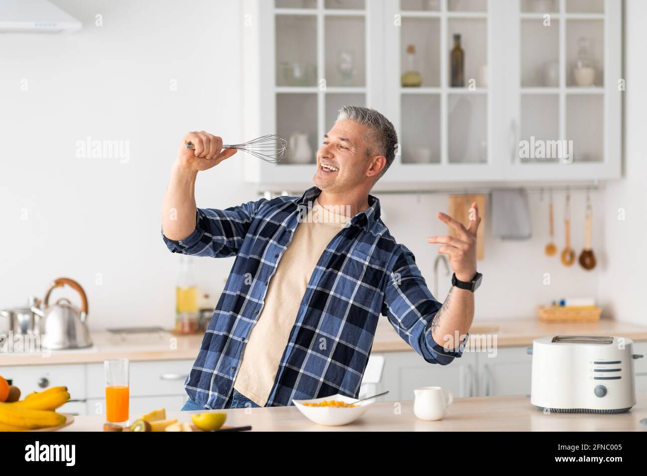 L'uomo anziano si fooling e cantando nel whisk mentre cucinando la prima colazione nell'interno moderno della cucina. Foto Stock