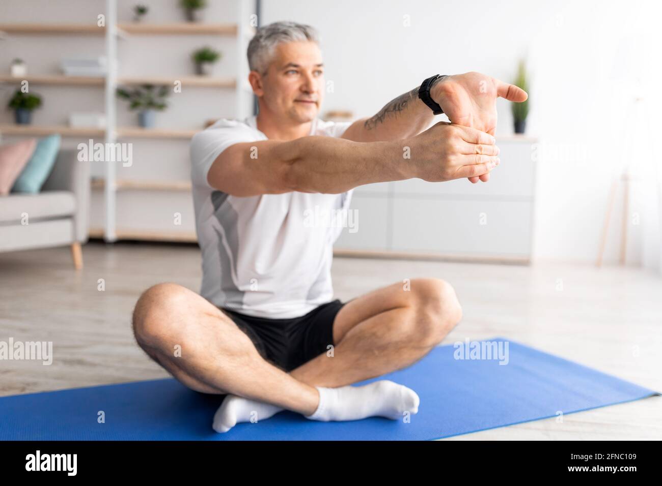 Uomo anziano sportivo che fa stretching per gli esercizi a mano, seduto sul tappetino nell'interno del soggiorno, spazio libero Foto Stock
