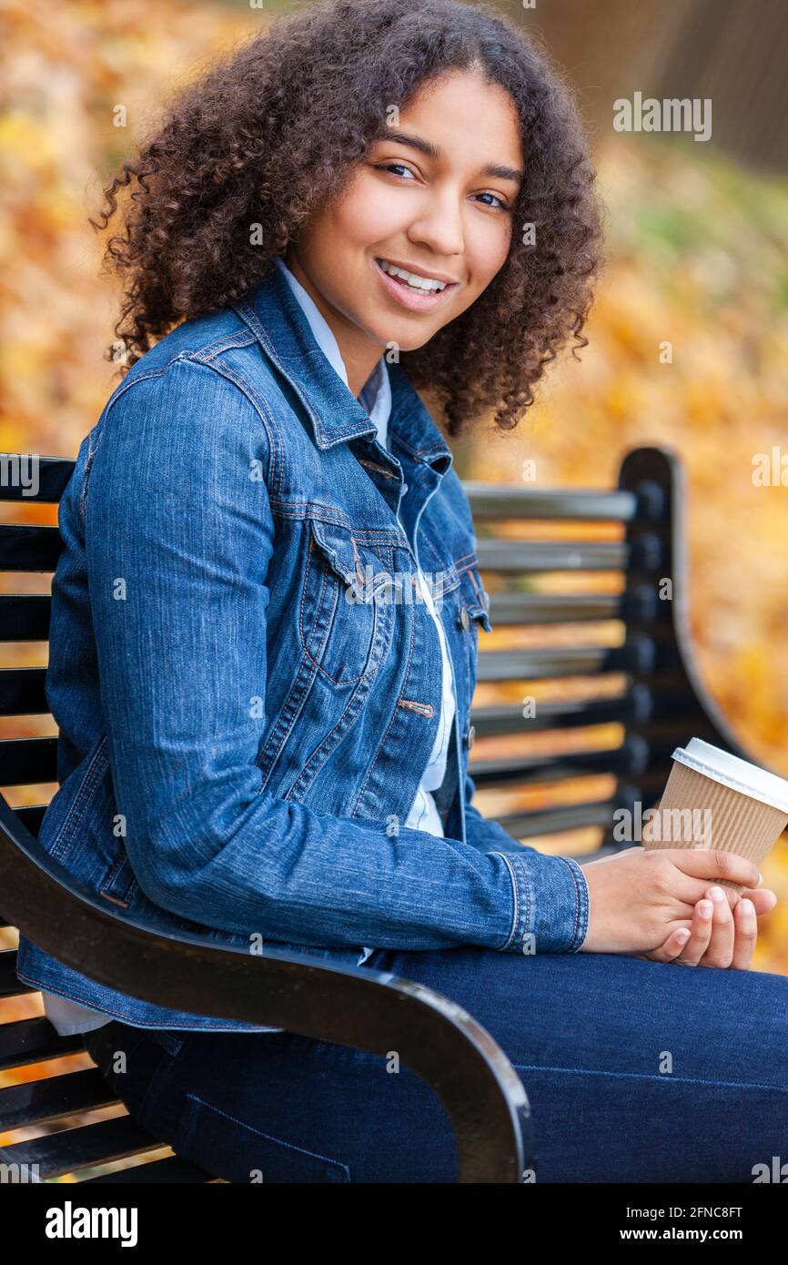 Bella felice mista razza Africana americana ragazza adolescente femmina giovane donna che sorride e beve caffè da asporto fuori seduta su un parcheggiare la panca in una Foto Stock