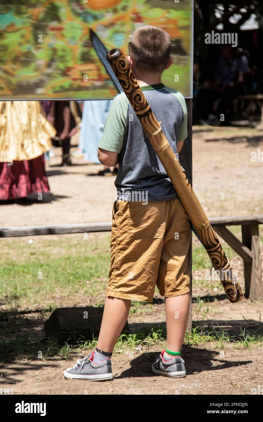 Ragazzo giovane che indossa pantaloncini e scarpe da tennis con didgeridoo  e. la spada di legno esamina la grande mappa in piedi al festival o fiera  con persone offuscate in termini di