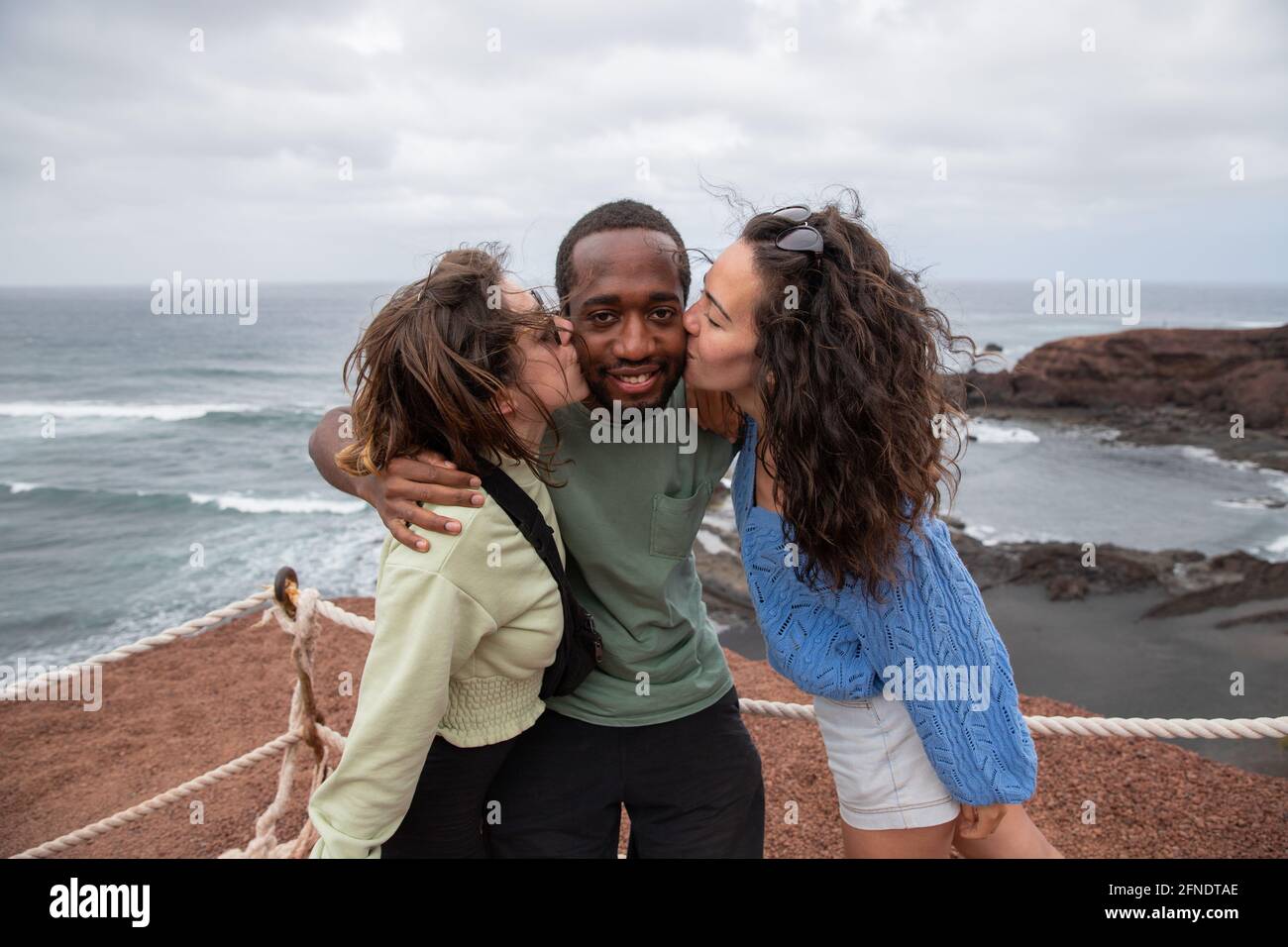 Un ragazzo africano riceve un bacio da due ragazze caucasiche durante una vacanza, un gruppo multirazziale felice sta avendo divertimento Foto Stock