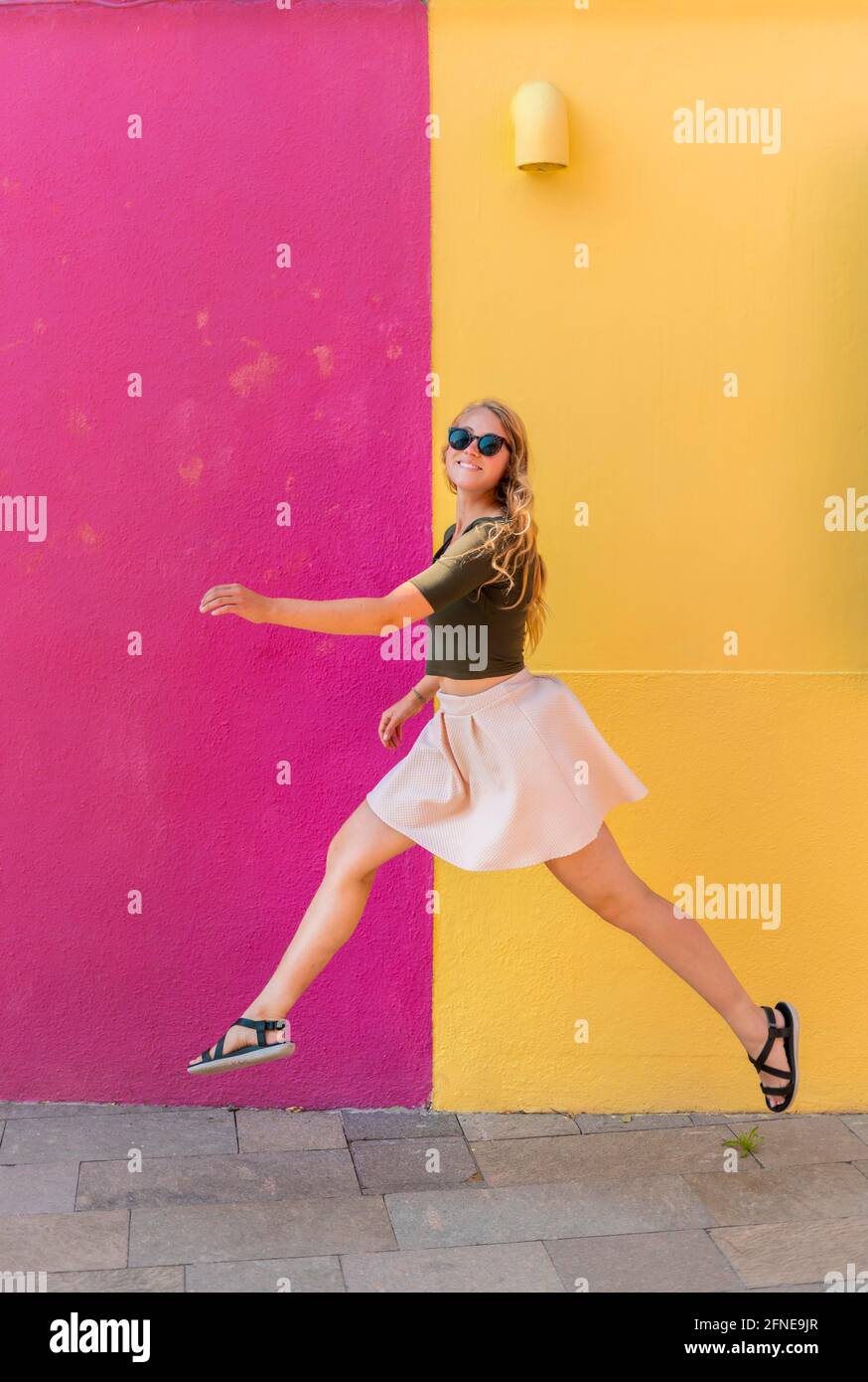 Giovane donna in abito salta felicemente di fronte a casa colorata, giallo e rosa facciata casa, Isola di Burano, Venezia, Veneto, Italia Foto Stock