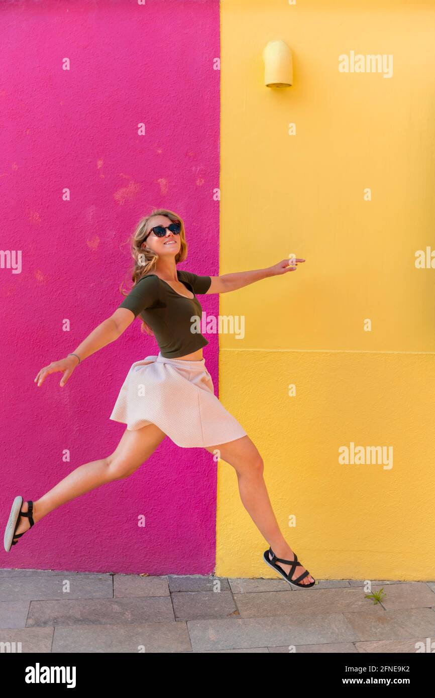 Giovane donna in abito salta felicemente di fronte a casa colorata, giallo e rosa facciata casa, Isola di Burano, Venezia, Veneto, Italia Foto Stock