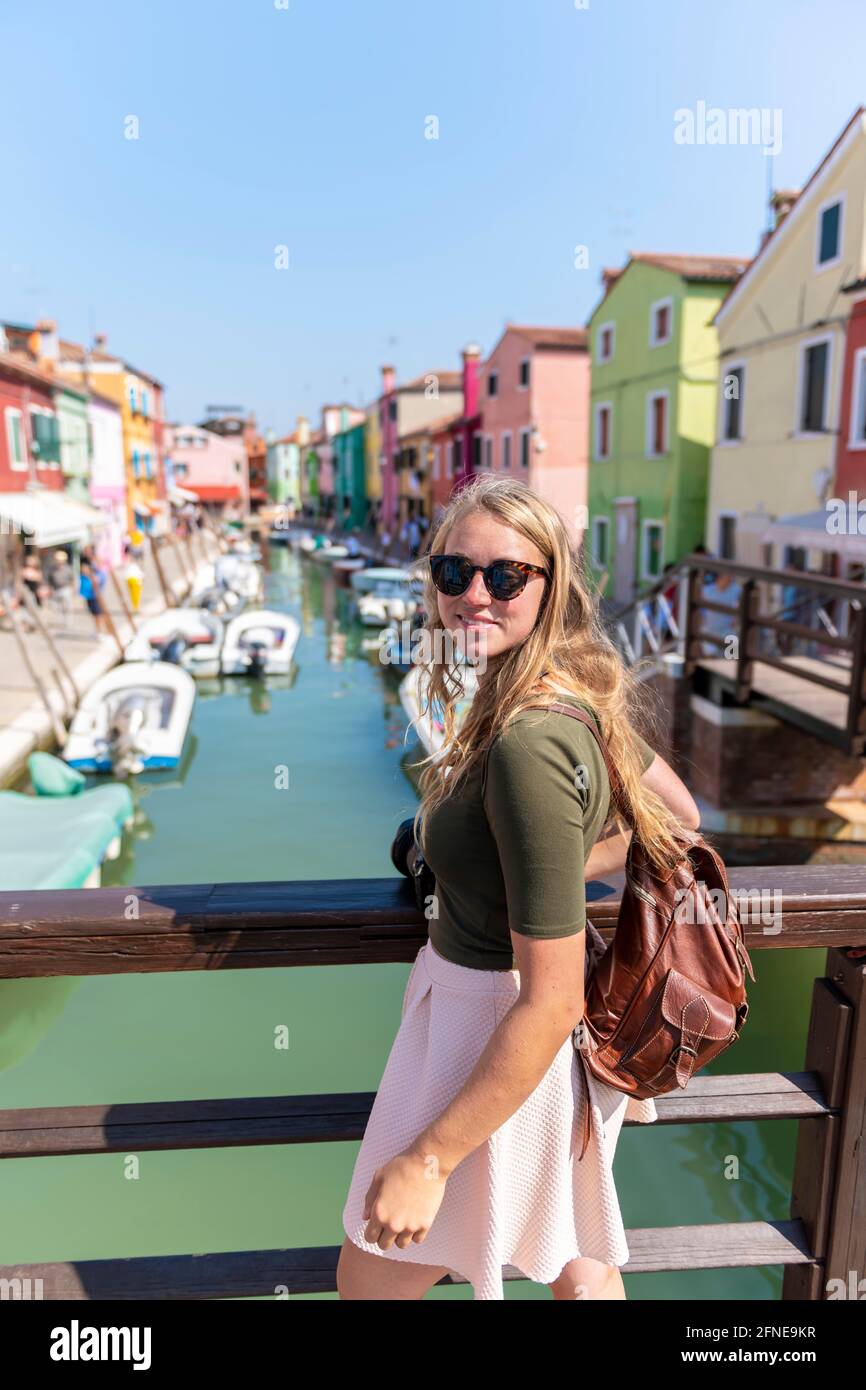 Giovane donna di fronte a case colorate, canale con barche e facciate colorate casa, Burano Island, Venezia, Veneto, Italia Foto Stock