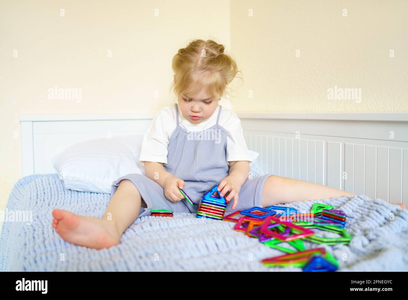 Bambina concentrata con capelli d'oro giocando con giocattolo educativo costruttore magnetico sul letto, piccolo bambino prescolare in abito carino godendo il tempo di gioco a casa. Concetto di sviluppo infantile Foto Stock