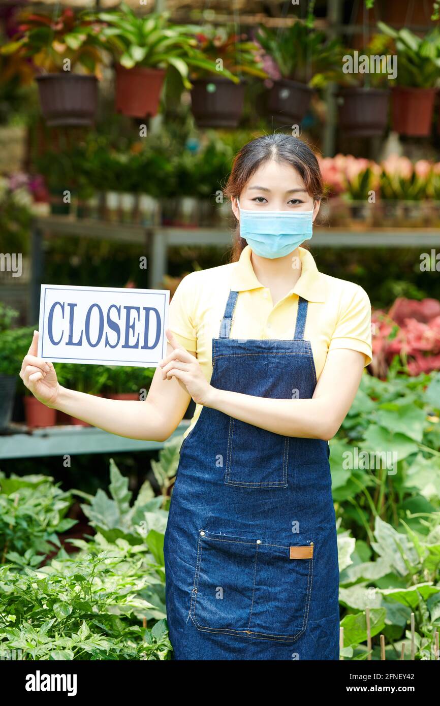 Bella giovane donna in maschera protettiva che chiude il centro di giardinaggio o. vivaio di piante a causa di pandemia di coronavirus Foto Stock