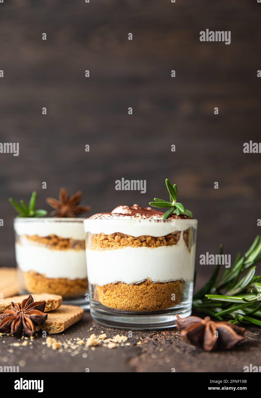 Dessert a strati in vaso di vetro con biscotti sbriciolati e panna montata decorata con rosmarino e anice, sfondo scuro. Non cuocere cheesecake, trifle o p Foto Stock