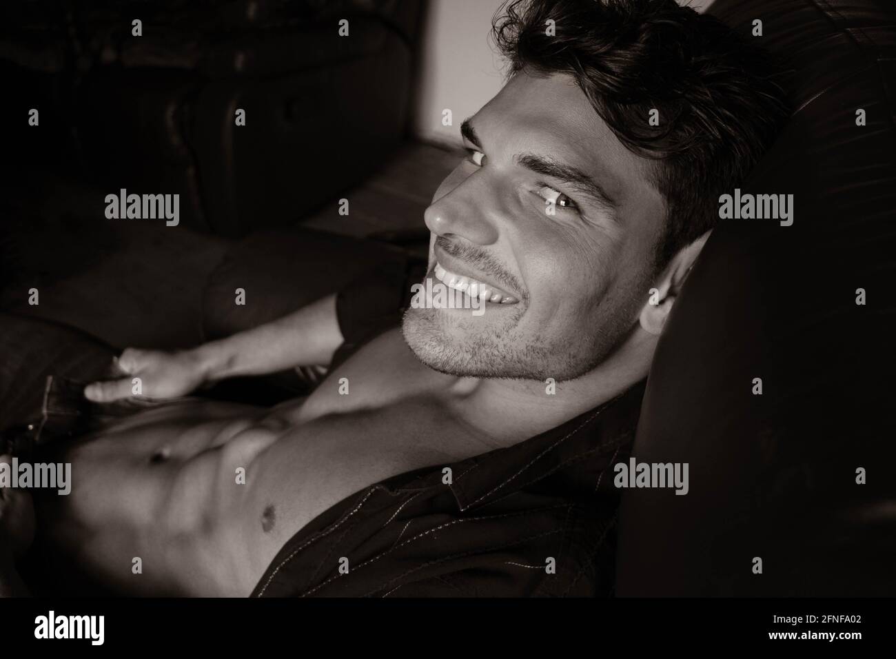 Uomo bello con la barba e camicia aperta rivelando abs muscolare seduti in poltrona in pelle guardando la fotocamera. Foto Stock