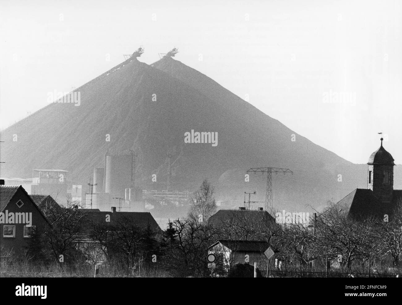 Alle spalle della città di Ronneburg sorgono i cumuli appuntiti di estrazione dell'uranio. La più grande area estrattiva di uranio in Europa si trova tra Gera e Karl-Marx-Stadt e sotto la gestione di SDAG, la società mista sovietico-tedesca. In primo piano si possono vedere le case del villaggio e qualche industria in background. Sulla destra si trova una chiesa. [traduzione automatizzata] Foto Stock