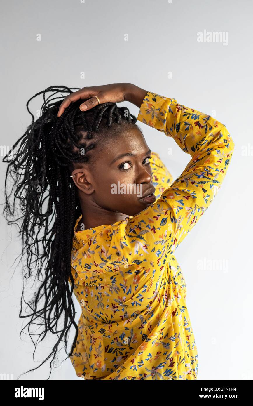 Vista laterale del modello femminile afro-americano con trecce a contatto capelli mentre si guarda la fotocamera su sfondo grigio in studio Foto Stock
